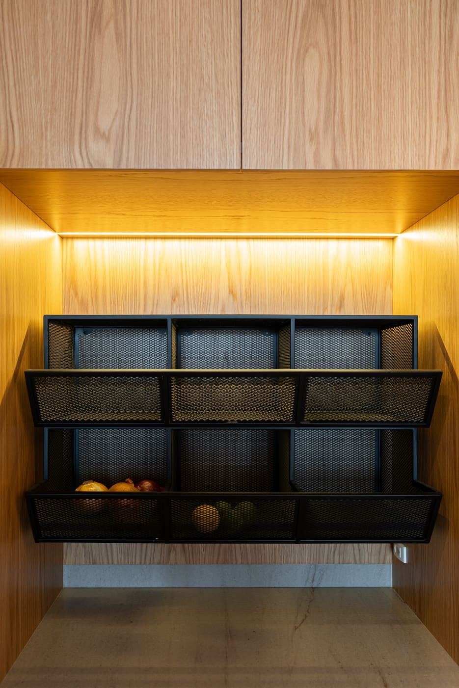 Lit pantry niche with black mesh fruit baskets set into wood cabinetry